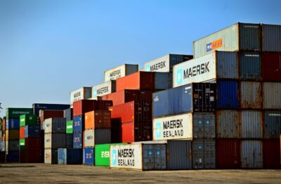 Stacked shipping containers at a busy port under a clear blue sky.