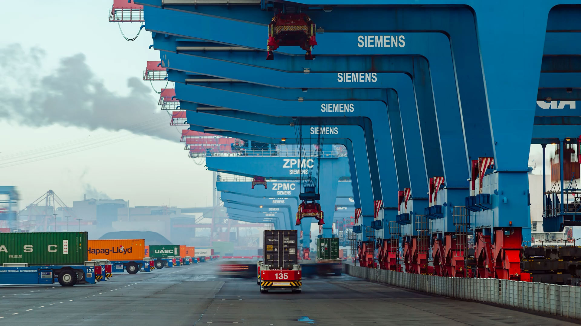Large blue cranes lined up at a shipping port with containers and vehicles.